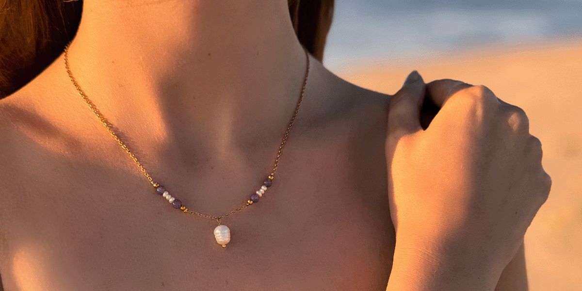 Une femme portant un collier doré avec une perle d'eau douce blanche et des pierres naturelles en améthyste, éclairée par la lumière d'un coucher de soleil.