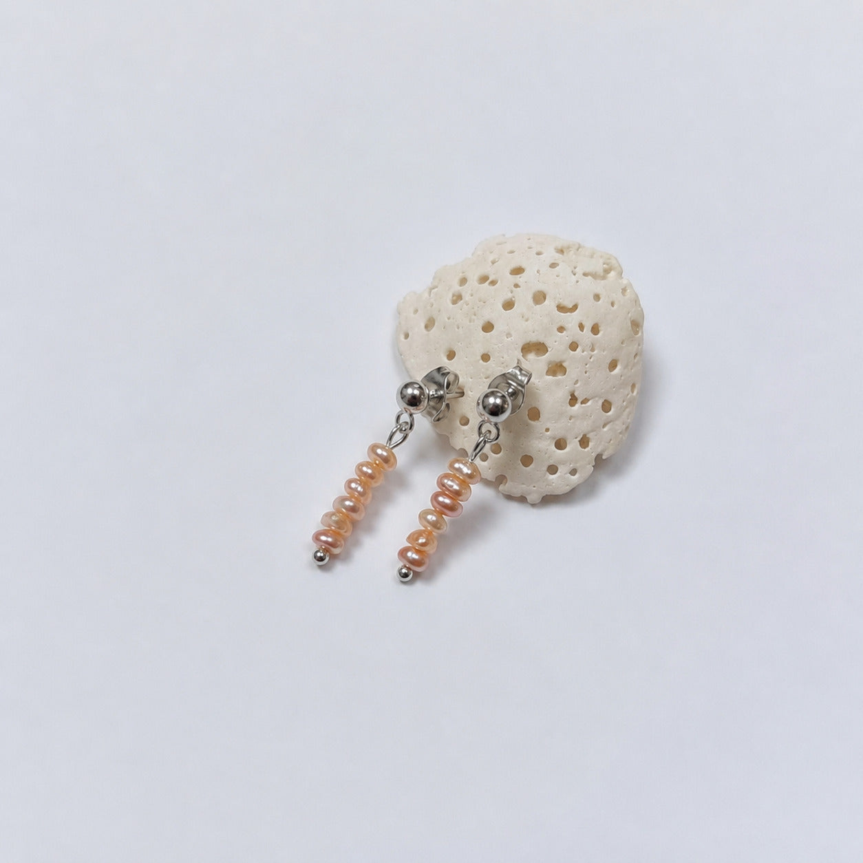 Paire de boucles d'oreilles puces argentées composées chacune d'une tige de quatre petites perles d'eau douce oranges empilées en dégradé de taille, posées contre un fragment de corail beige poreux sur fond gris clair.