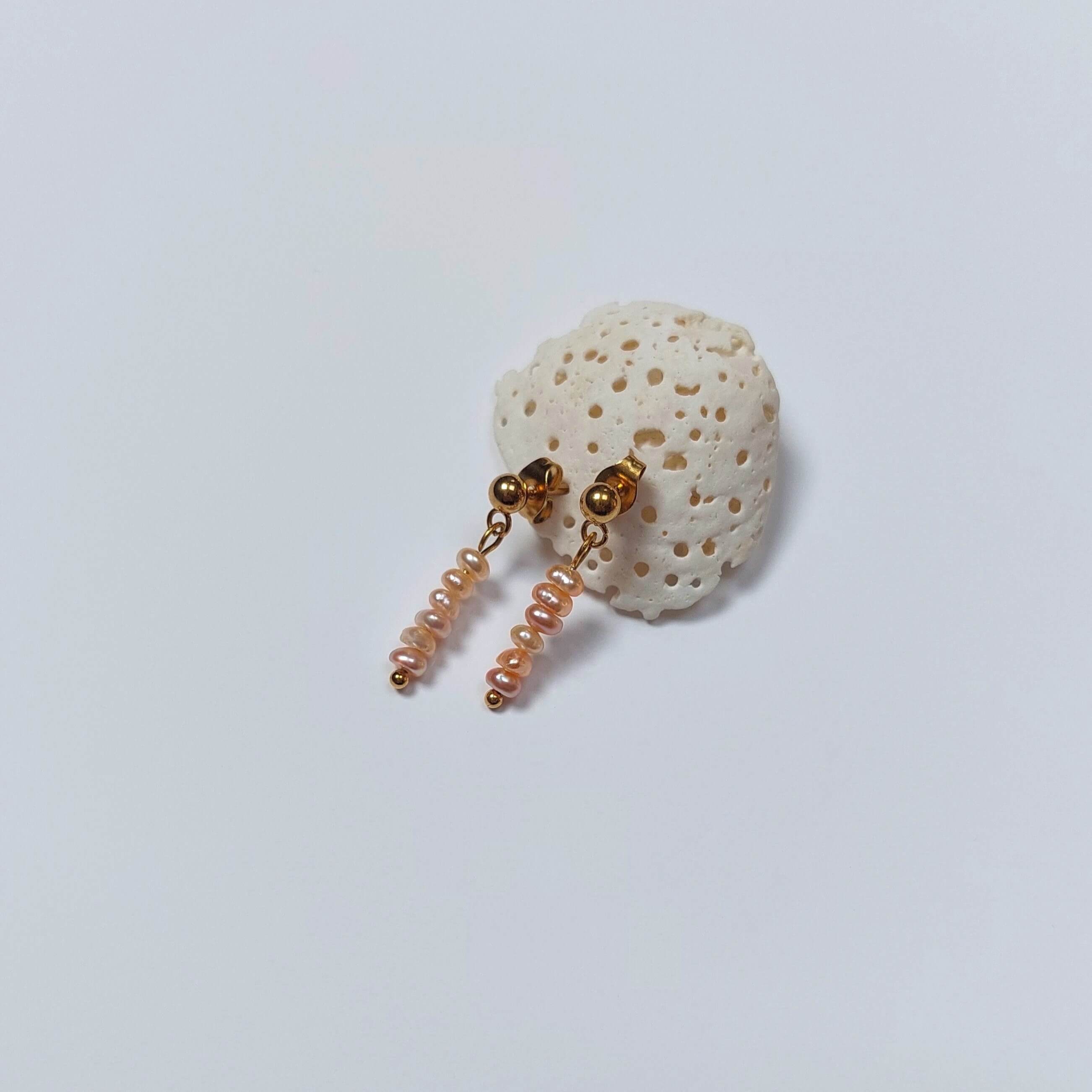 Boucles d’oreilles pendantes en perles d’eau douce naturelles oranges et acier inoxydable doré, au design linéaire et contemporain, accrochées à morceau de corail blanc, sur fond neutre.