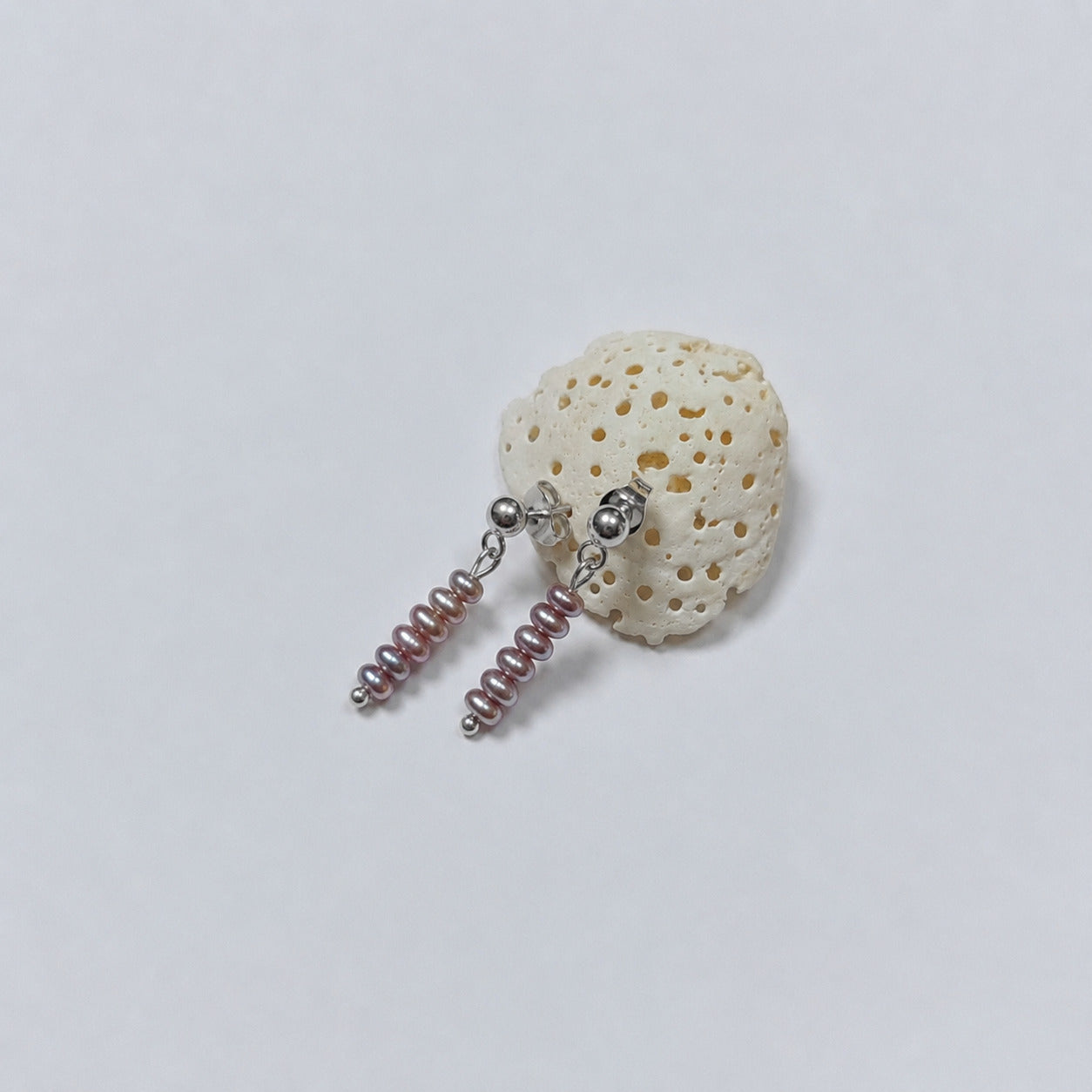 Paire de boucles d'oreilles puces argentées composées chacune d'une tige de quatre petites perles d'eau douce violettes empilées en dégradé de taille, posées contre un fragment de corail beige poreux sur fond gris clair.