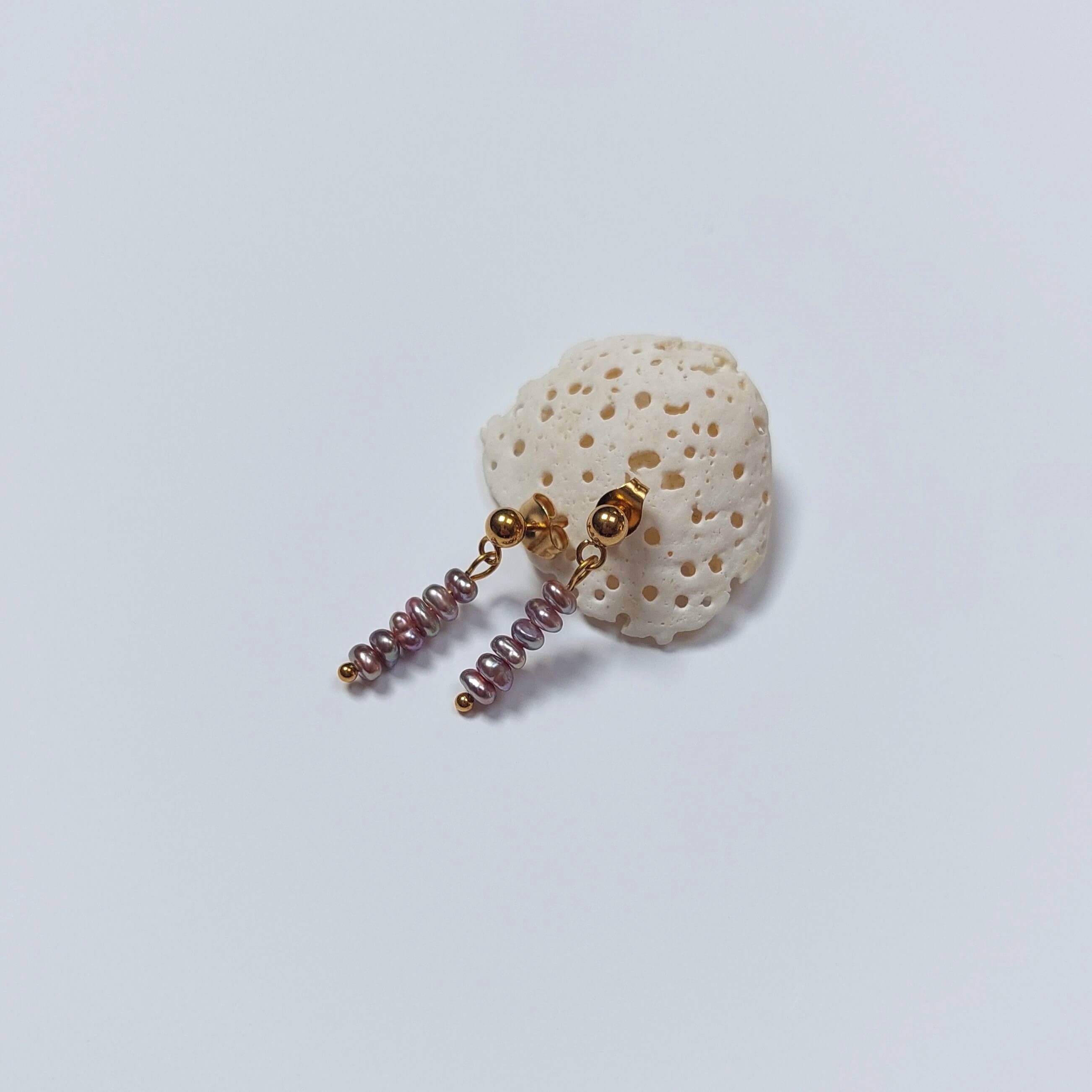 Boucles d’oreilles pendantes en perles d’eau douce naturelles violettes et acier inoxydable doré, au design linéaire et contemporain, accrochées à morceau de corail blanc, sur fond neutre.