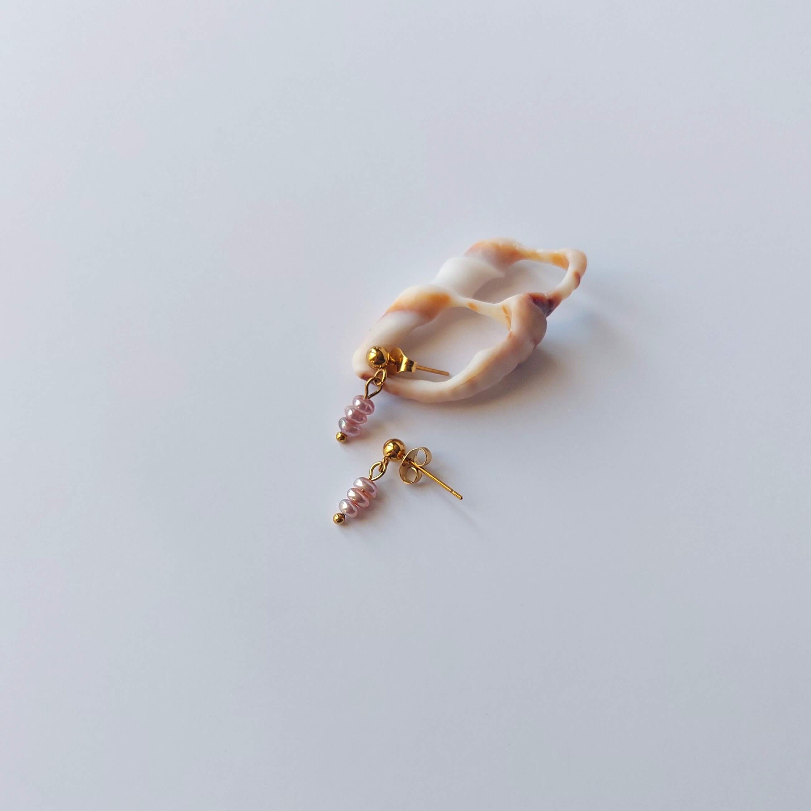 Boucles d’oreilles pendantes en acier inoxydable doré avec perles d’eau douce violettes, posées sur un coquillage, sur un fond neutre.