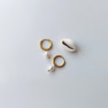 Boucles d’oreilles créoles en acier inoxydable doré avec une perle d’eau douce naturelle blanche, posées à côté d’un coquillage, sur un fond neutre. 