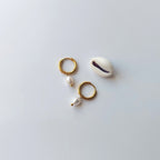 Boucles d’oreilles créoles en acier inoxydable doré avec une perle d’eau douce naturelle blanche, posées à côté d’un coquillage, sur un fond neutre. 
