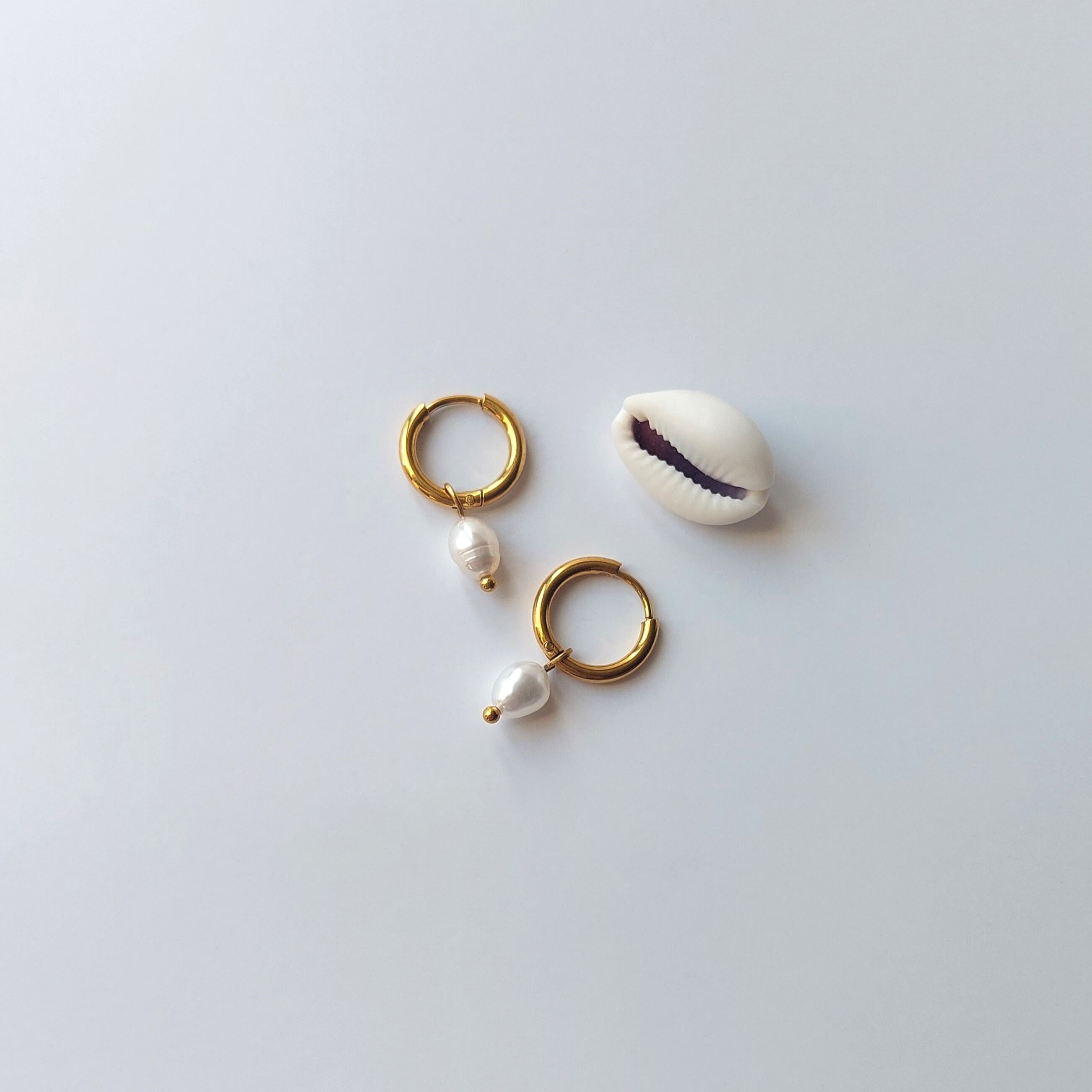 Boucles d’oreilles créoles en acier inoxydable doré avec une perle d’eau douce naturelle blanche, posées à côté d’un coquillage, sur un fond neutre. 