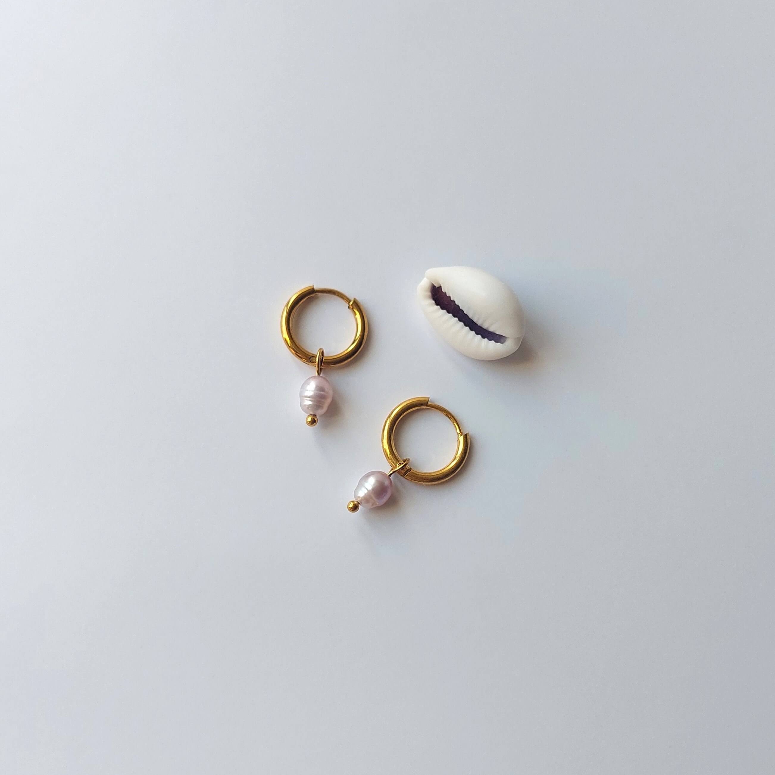 Boucles d’oreilles créoles en acier inoxydable doré avec une perle d’eau douce naturelle violette, posées à côté d’un coquillage, sur un fond neutre. 