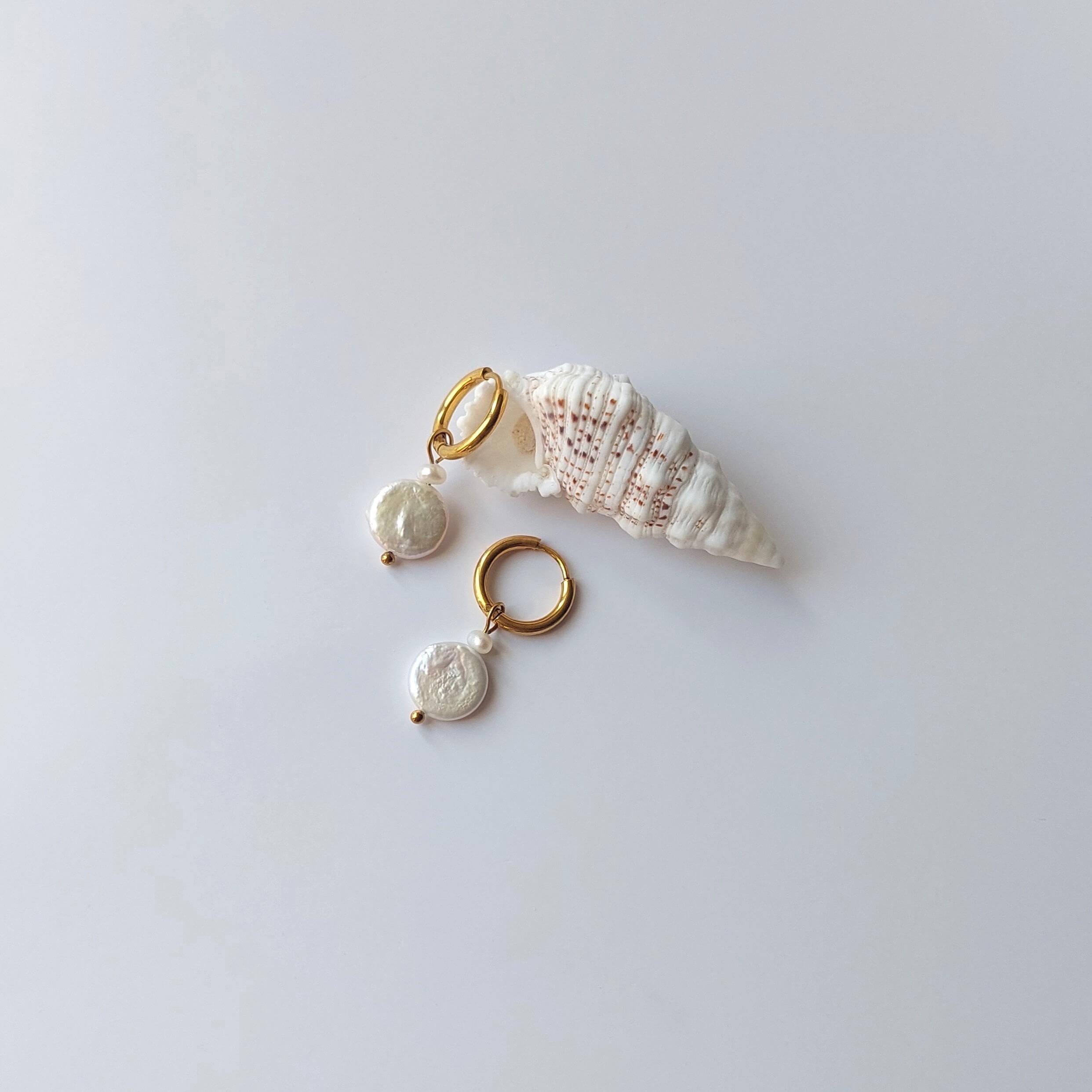 Boucles d’oreilles créoles en acier inoxydable doré avec perles d’eau douce naturelles blanches, posées sur un coquillage, style élégant, sur fond neutre.
