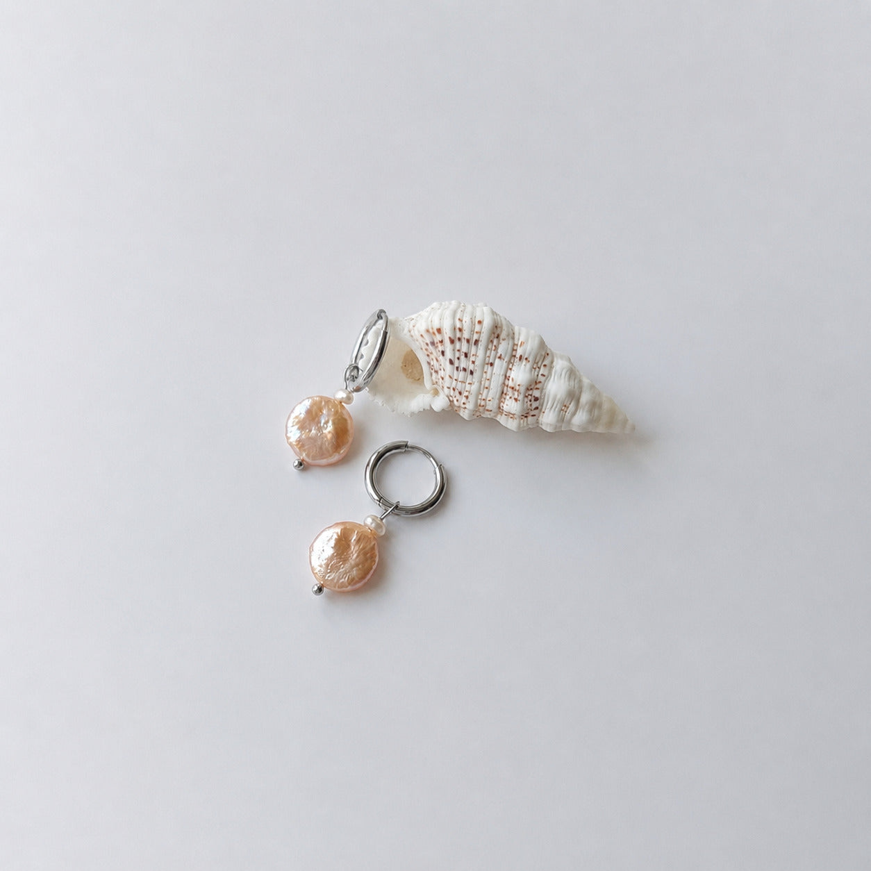 Paire de boucles d'oreilles créoles argentées ornées chacune d'une petite perle de rocaille et d'une grande perle d'eau douce plate ronde nacrée en pendentif, posées sur fond gris clair aux côtés d'un coquillage en spirale blanc et beige.