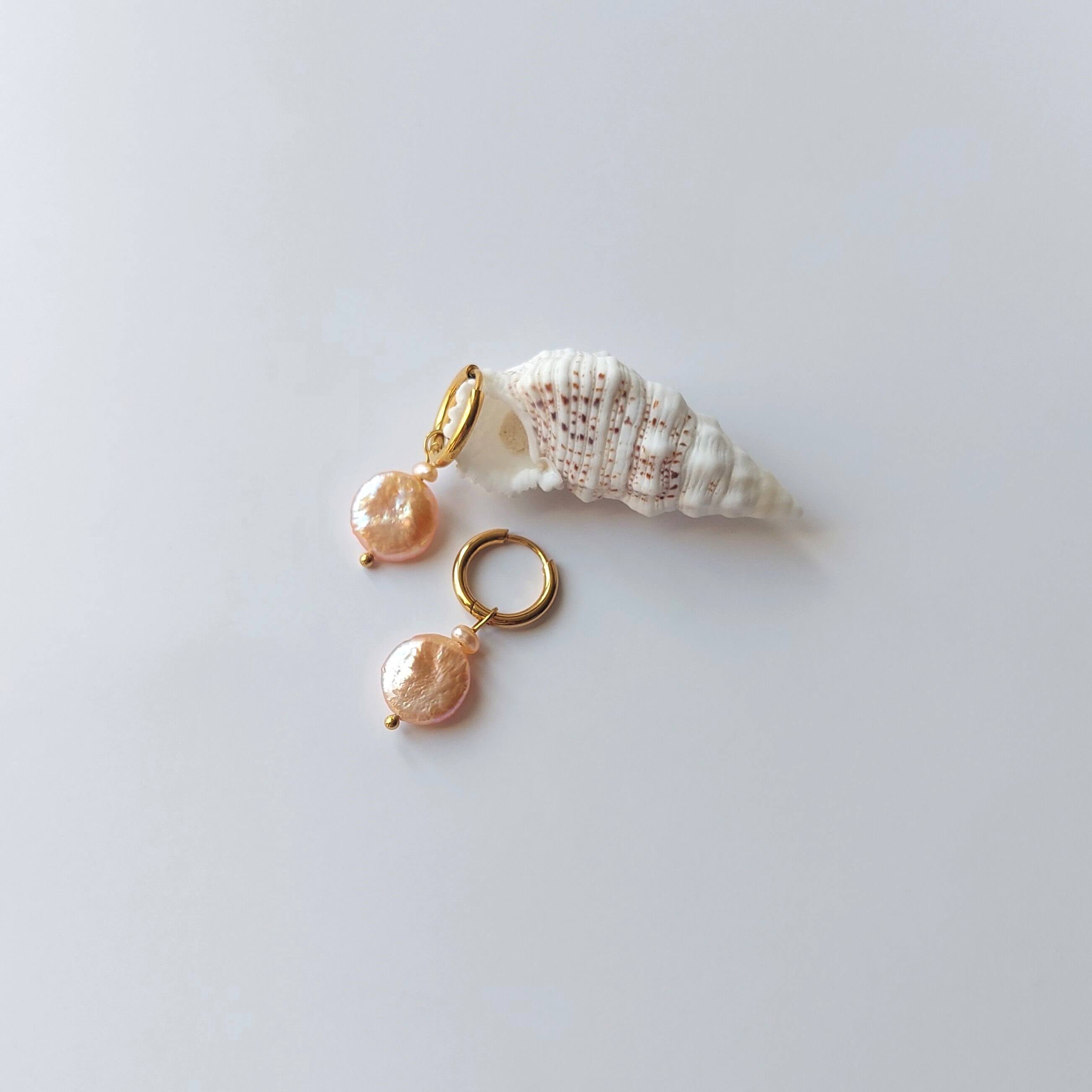Boucles d’oreilles créoles en acier inoxydable doré avec perles d’eau douce naturelles oranges, posées sur un coquillage, style élégant, sur fond neutre.