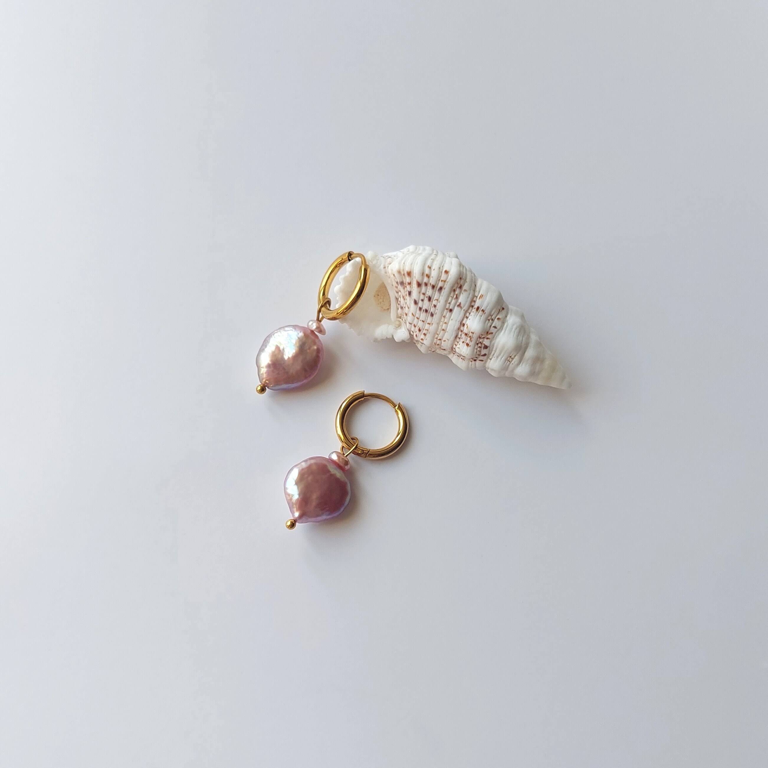 Boucles d’oreilles créoles en acier inoxydable doré avec perles d’eau douce naturelles violettes, posées sur un coquillage, style élégant, sur fond neutre.