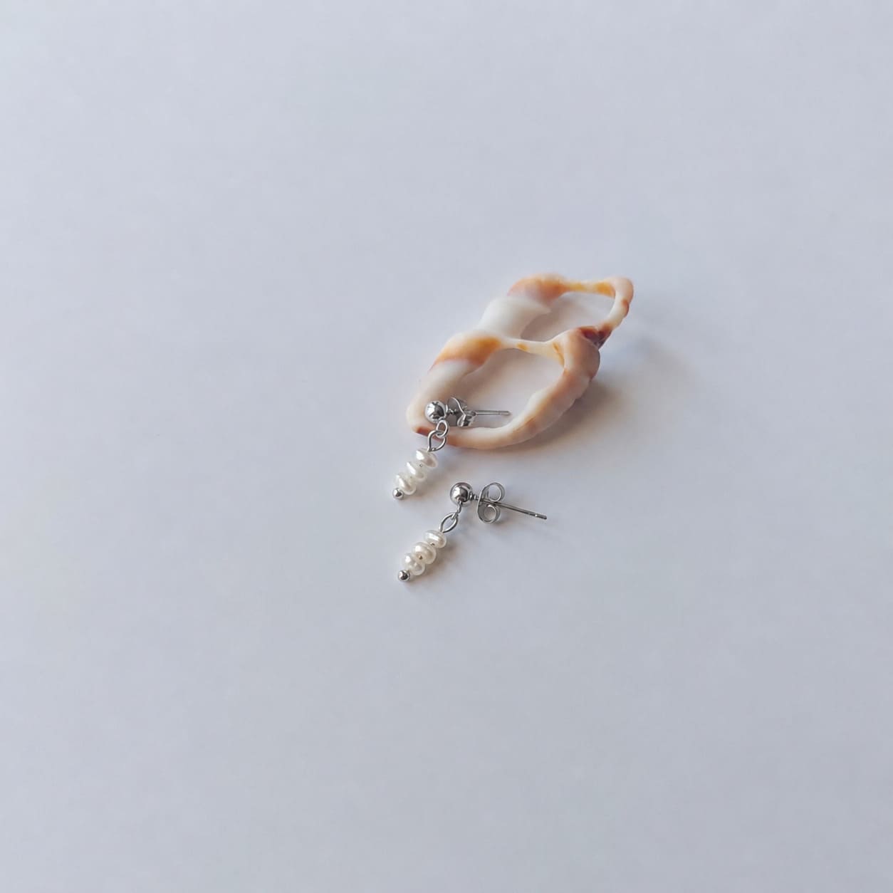 Boucles d’oreilles pendantes en acier inoxydable argenté avec perles d’eau douce blanches, posées sur un coquillage, sur un fond neutre.