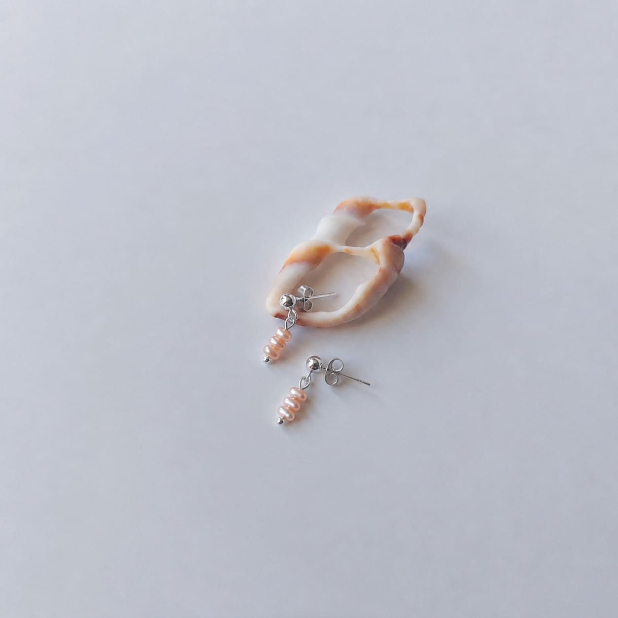 Boucles d’oreilles pendantes en acier inoxydable argenté avec perles d’eau douce oranges, posées sur un coquillage, sur un fond neutre.