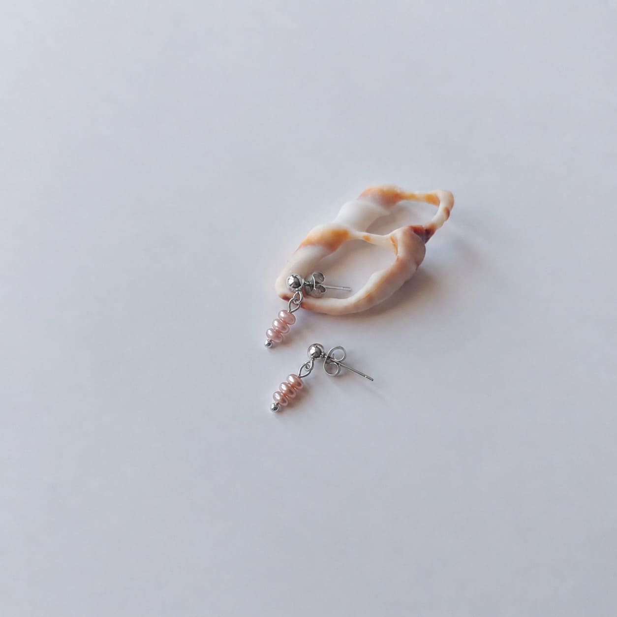 Boucles d’oreilles pendantes en acier inoxydable argenté avec perles d’eau douce violettes, posées sur un coquillage, sur un fond neutre.