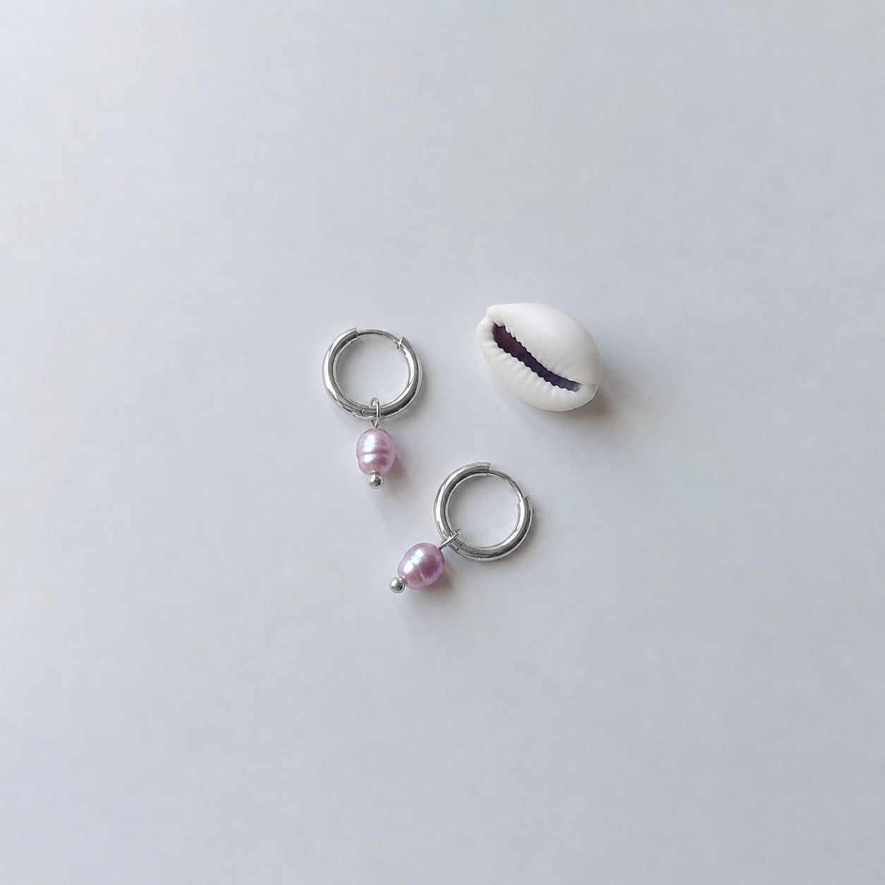 Paire de boucles d'oreilles créoles argentées ornées chacune d'une perle d'eau douce violette baroque en pendentif, posées sur fond gris clair aux côtés d'un coquillage cauri blanc.