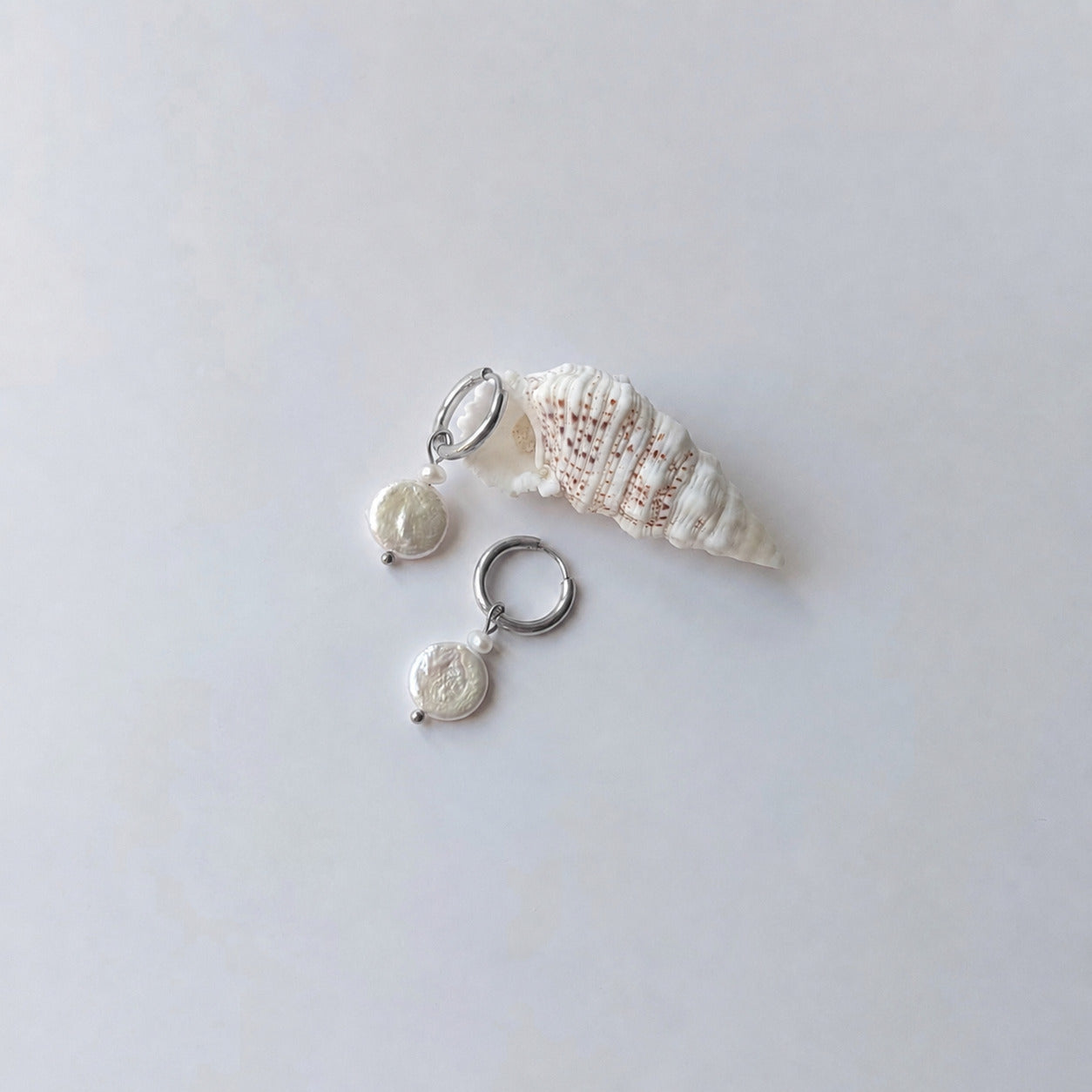 Paire de boucles d'oreilles créoles argentées ornées chacune d'une petite perle de rocaille et d'une grande perle d'eau douce plate ronde nacrée en pendentif, posées sur fond gris clair aux côtés d'un coquillage en spirale blanc et beige.