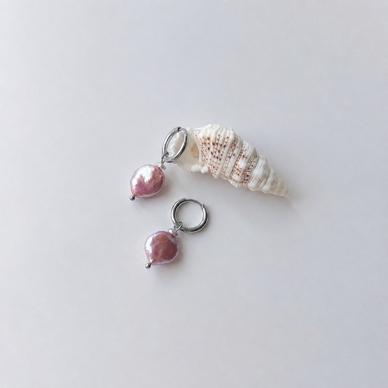 Paire de boucles d'oreilles créoles argentées ornées chacune d'une petite perle de rocaille et d'une grande perle d'eau douce plate ronde nacrée en pendentif, posées sur fond gris clair aux côtés d'un coquillage en spirale blanc et beige.