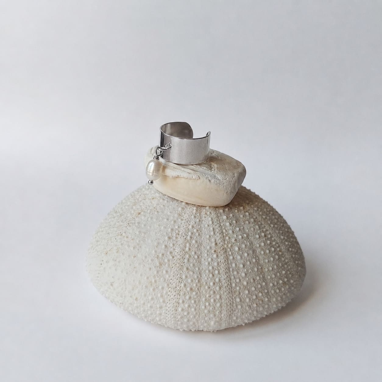 Bague chevalière large argentée ouverte ornée d'une perle d'eau douce ovale de couleur blanche en pendentif, posée sur un coquillage blanc déposé sur un oursin séché, fond gris clair.