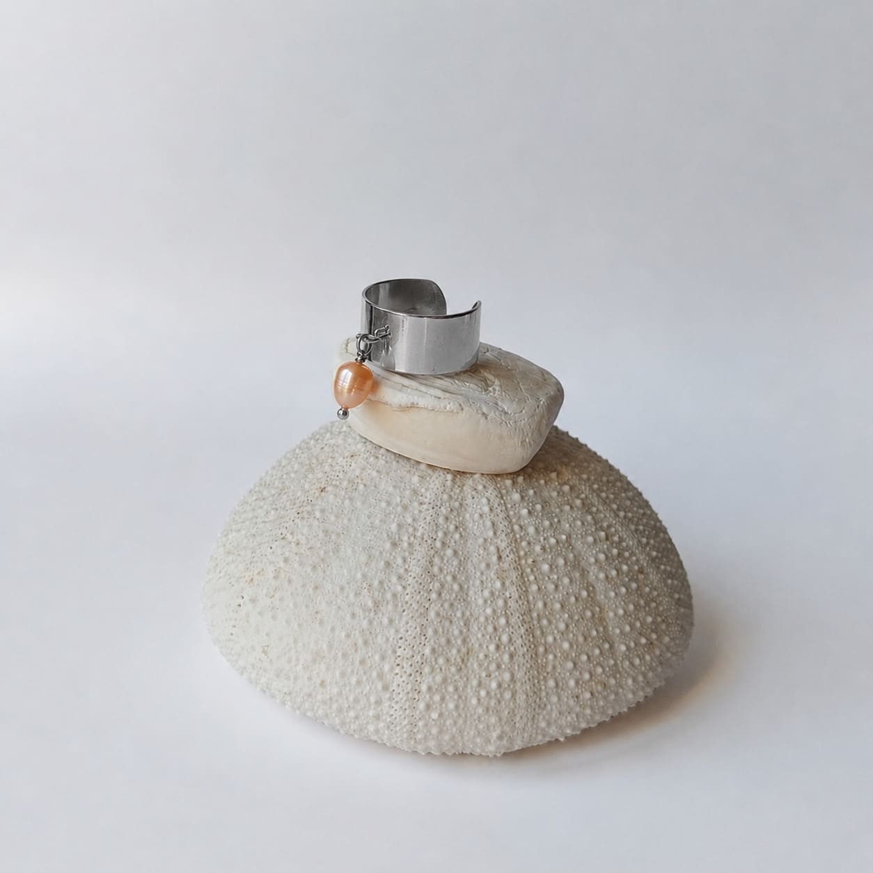 Bague chevalière large argentée ouverte ornée d'une perle d'eau douce ovale de couleur pêche-orange en pendentif, posée sur un coquillage blanc déposé sur un oursin séché, fond gris clair.