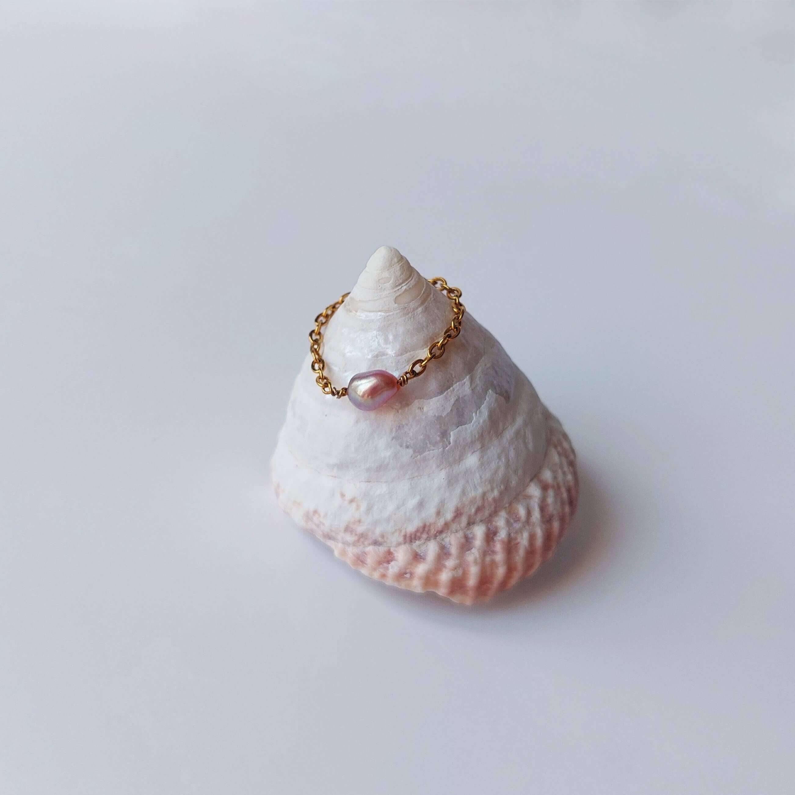 Bague faite d'une chaine fine en acier inoxydable doré et perle d'eau douce violette posée sur un coquillage