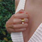 Femme portant la bague Pavona ajustable en acier inoxydable doré, large anneau lisse orné de breloques perle d'eau douce baroque et pierre de quartz fraise rose, photographiée en extérieur avec un haut en crochet blanc et un fond de végétation verte.