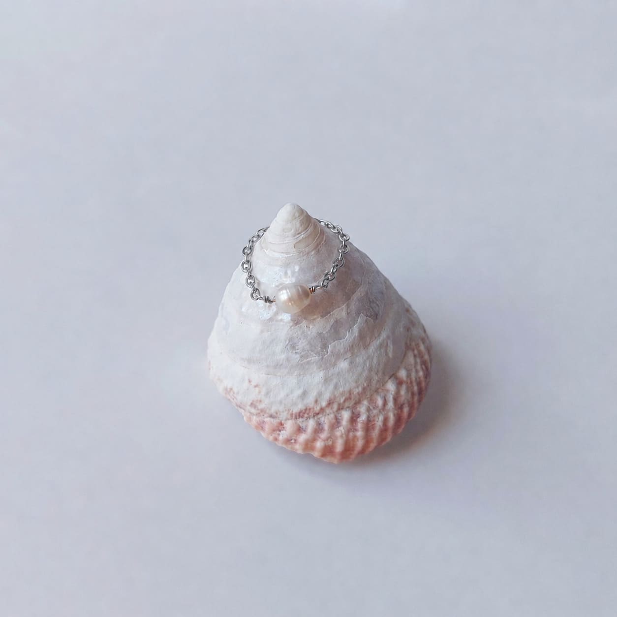 Bague fine sur chaînette argentée ornée d'une unique perle d'eau douce blanche baroque, posée en équilibre sur un coquillage en spirale blanc et rose sur fond gris clair.