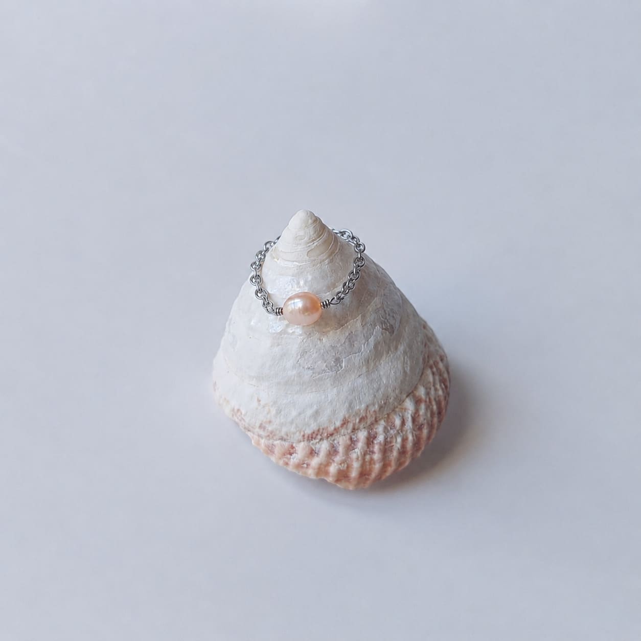 Bague fine sur chaînette argentée ornée d'une unique perle d'eau douce orange baroque, posée en équilibre sur un coquillage en spirale blanc et rose sur fond gris clair.