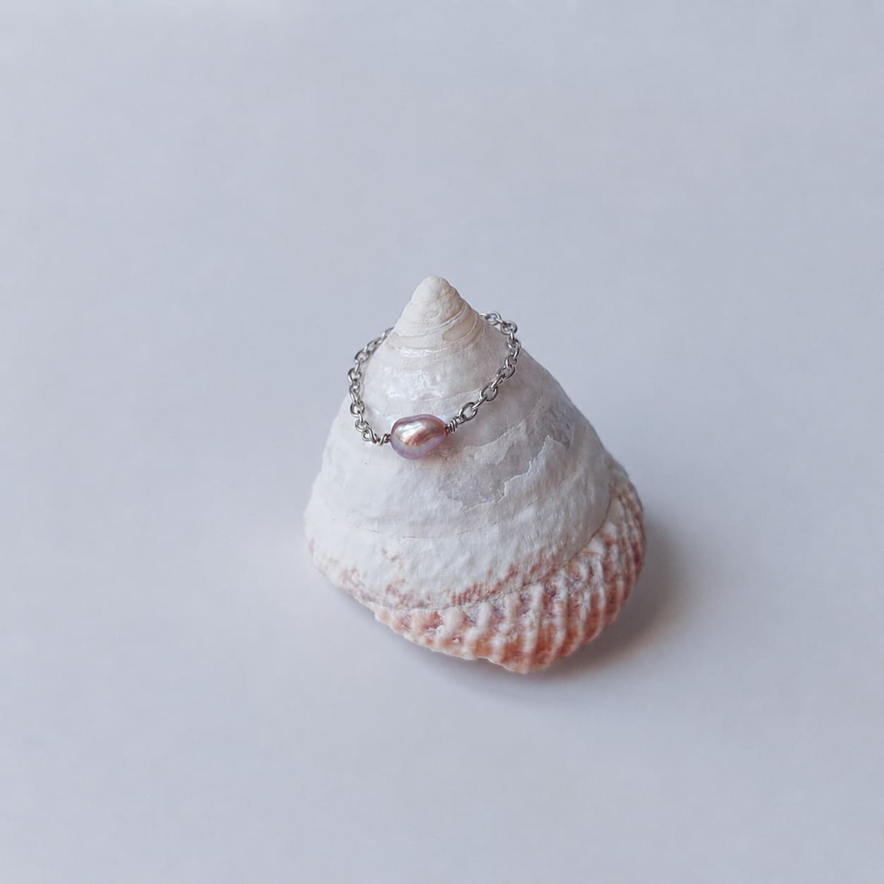 Bague fine sur chaînette argentée ornée d'une unique perle d'eau douce violette baroque, posée en équilibre sur un coquillage en spirale blanc et rose sur fond gris clair.