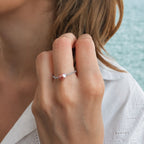 Bague fine sur chaînette argentée torsadée ornée d'une unique perle d'eau douce violette-mauve baroque, portée à l'annulaire par une femme en chemise blanche brodée avec un fond de mer turquoise en bokeh.