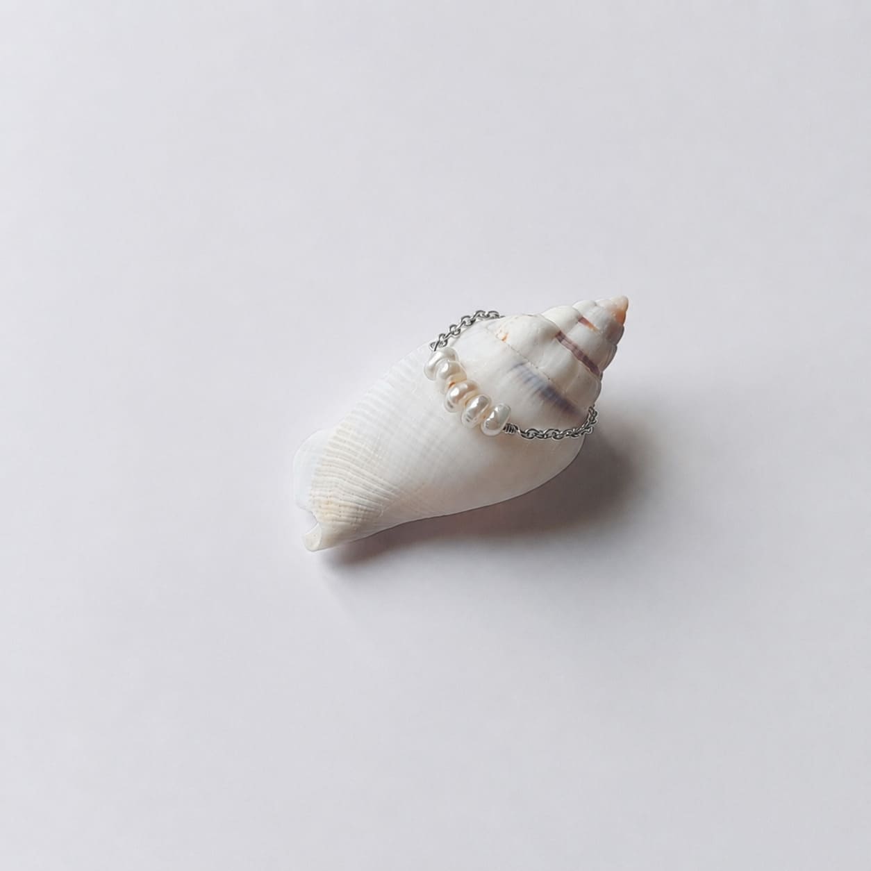 Bague fine sur chaînette argentée ornée de quatre petites perles d'eau douce blanches alignées, posée en équilibre sur un coquillage en spirale blanc et beige rayé sur fond gris clair.