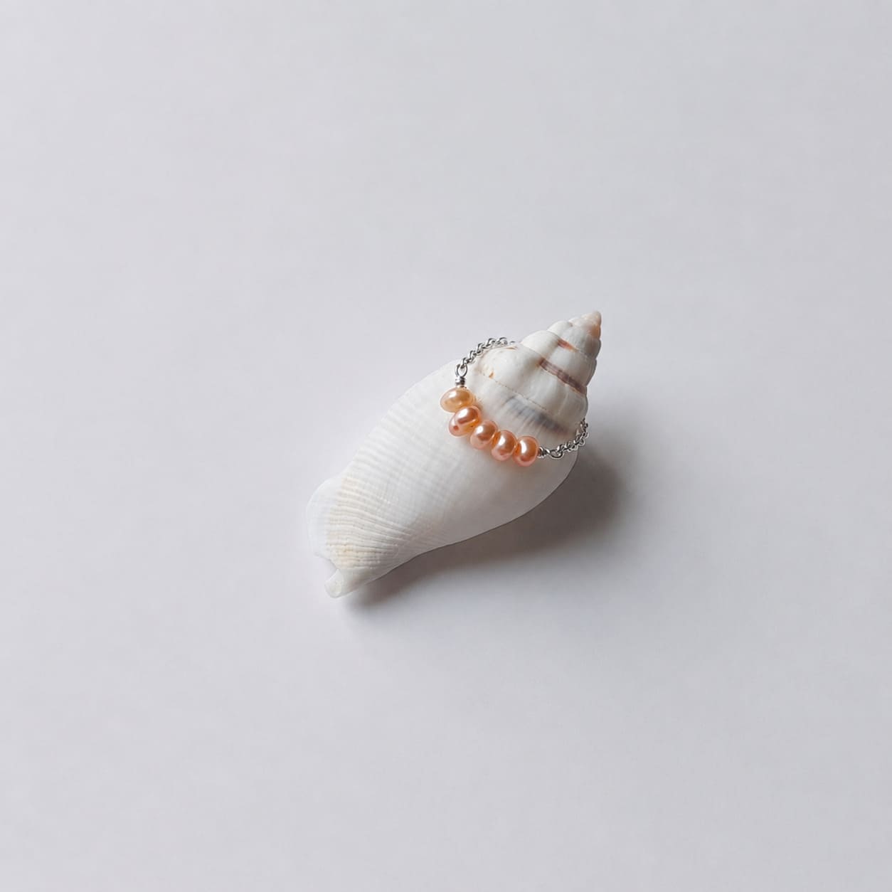 Bague fine sur chaînette argentée ornée de quatre petites perles d'eau douce oranges alignées, posée en équilibre sur un coquillage en spirale blanc et beige rayé sur fond gris clair.