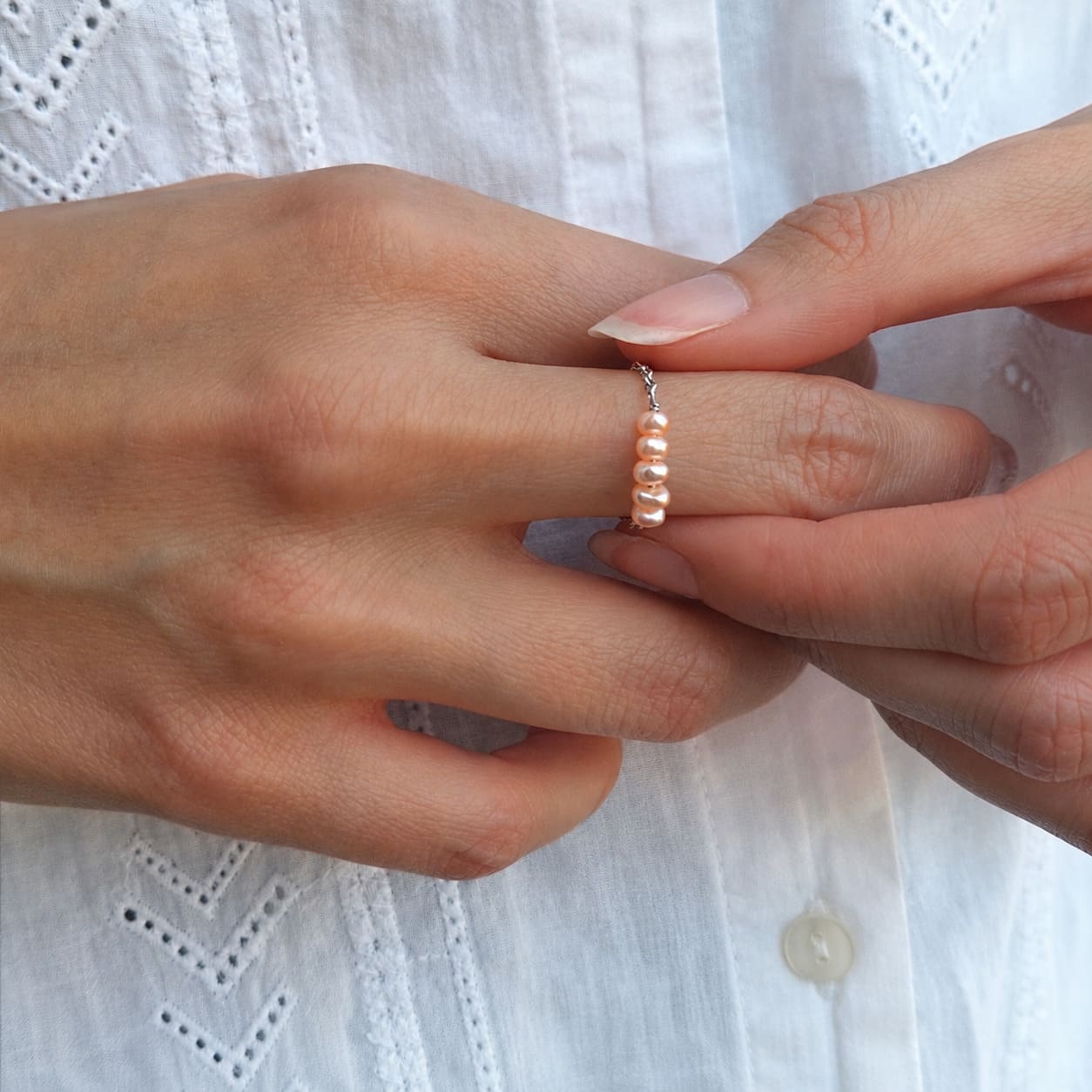 Bague fine sur chaînette argentée ornée de quatre petites perles d'eau douce pêche-orange alignées, portée à l'annulaire par une femme aux ongles naturels, vêtue d'une chemise blanche brodée.