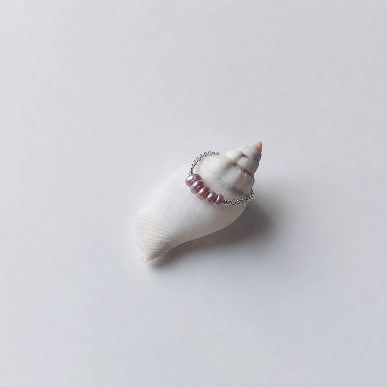 Bague fine sur chaînette argentée ornée de quatre petites perles d'eau douce violettes-mauves alignées, posée en équilibre sur un coquillage en spirale blanc et beige rayé sur fond gris clair.