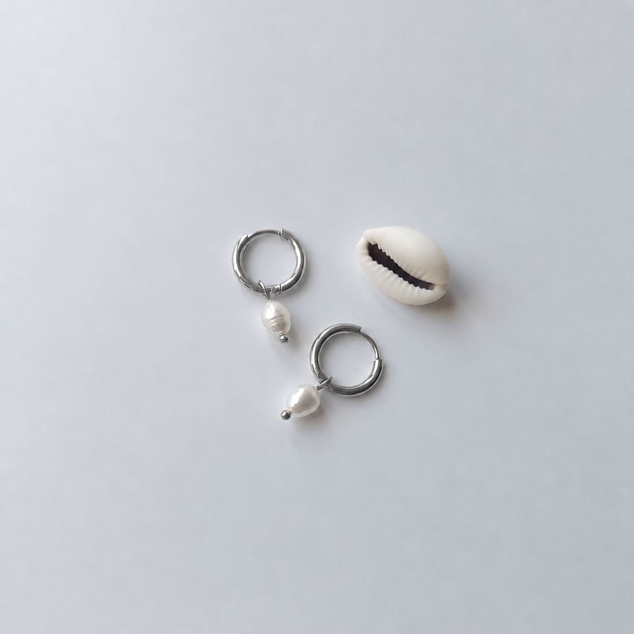 Paire de boucles d'oreilles créoles argentées ornées chacune d'une perle d'eau douce blanche baroque en pendentif, posées sur fond gris clair aux côtés d'un coquillage blanc.