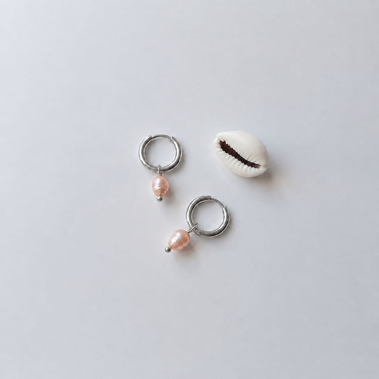Paire de boucles d'oreilles créoles argentées ornées chacune d'une perle d'eau douce orange baroque en pendentif, posées sur fond gris clair aux côtés d'un coquillage cauri blanc.