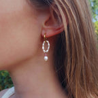 Femme portant la boucle d'oreille Polyphyllia en créole dorée en acier inoxydable avec un anneau de petites perles d'eau douce, une pierre de quartz fraise rose et une perle d'eau douce baroque en pendentif, photographiée en extérieur avec un fond de végétation verte.