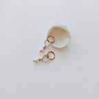 Paire de boucles d'oreilles Polyphyllia en créoles dorées en acier inoxydable avec un anneau de petites perles d'eau douce, une pierre de quartz fraise rose et une perle d'eau douce baroque en pendentif, posées contre un grand coquillage blanc nacré sur fond gris clair.