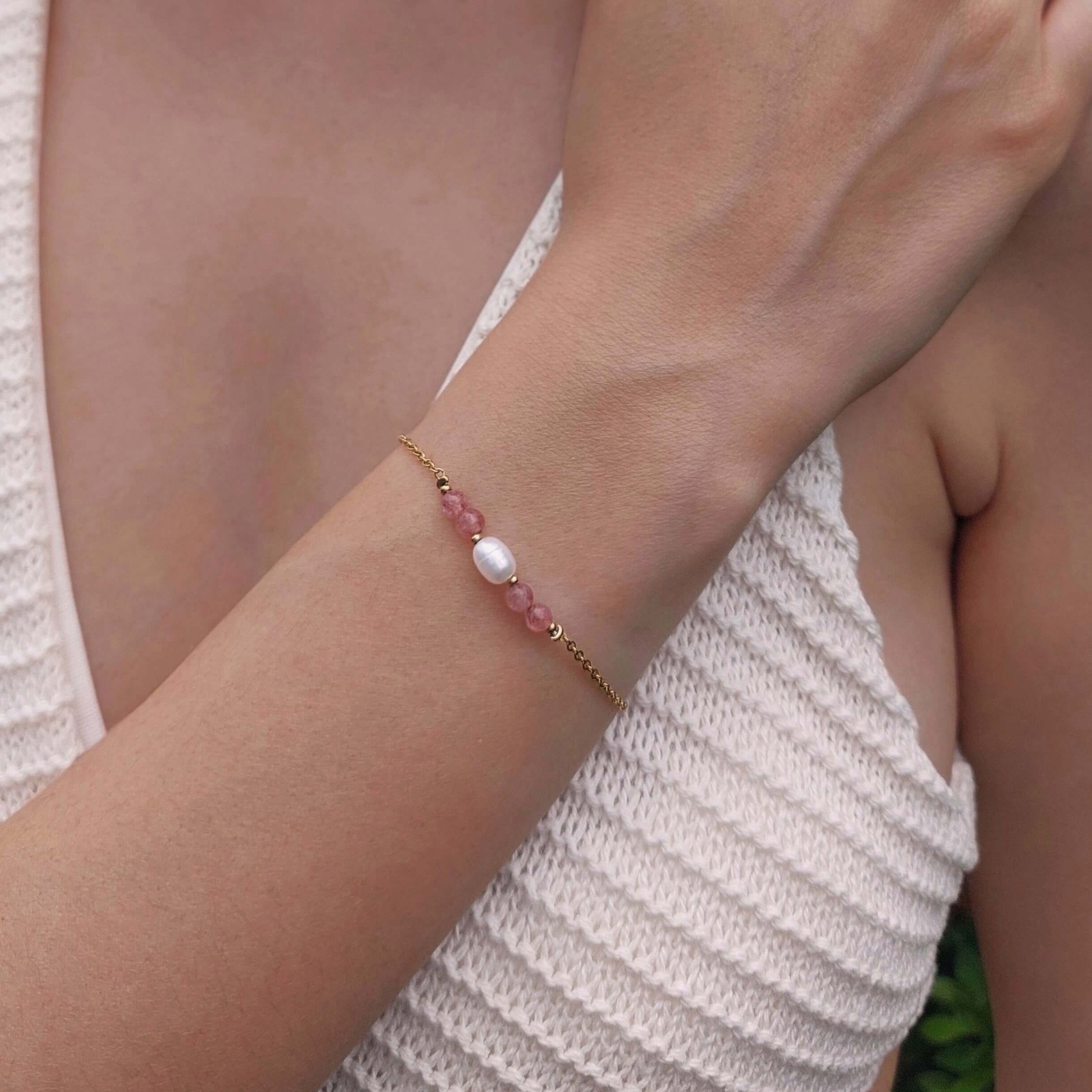 Femme portant le bracelet Acropora en quartz fraise et perle d'eau douce baroque centrale sur fine chaîne dorée en acier inoxydable, photographiée en extérieur avec un haut en crochet blanc et un fond de végétation verte.