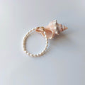 Bracelet en acier inoxydable doré orné de perles d’eau douce naturelles blanches, posé devant un coquillage rose, sur fond neutre.