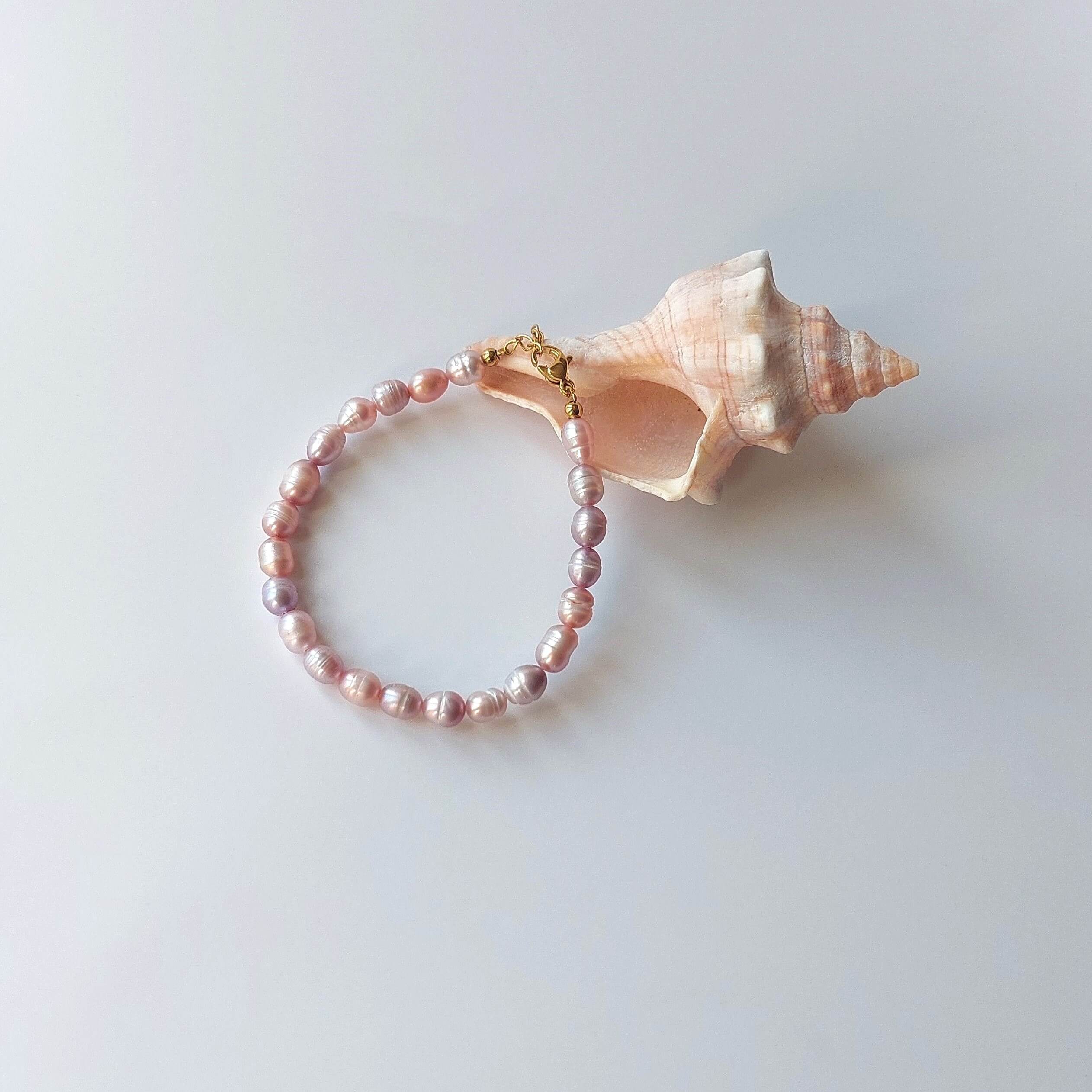Bracelet en acier inoxydable doré orné de perles d’eau douce naturelles violettes, posé devant un coquillage rose, sur fond neutre.