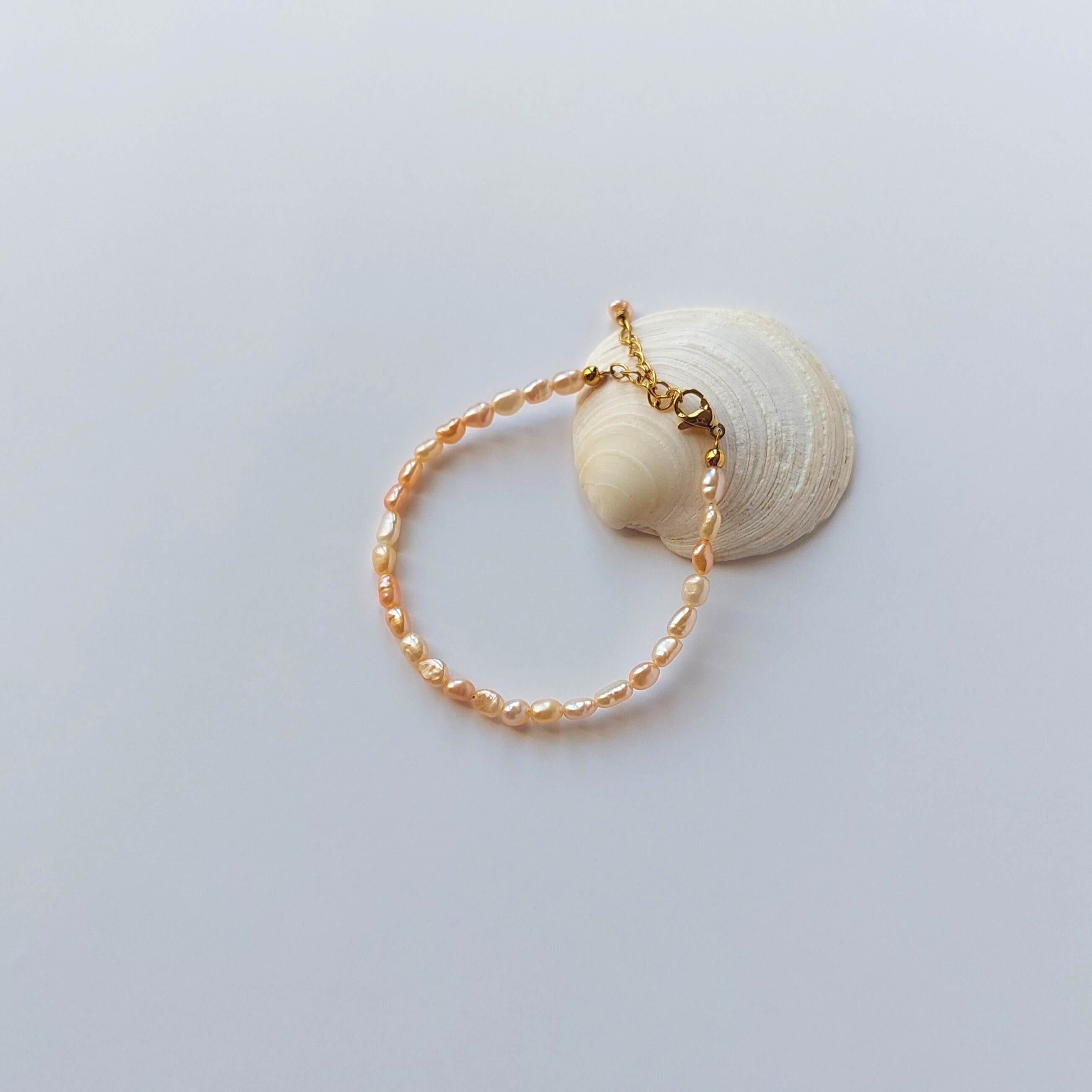 Bracelet en acier inoxydable doré orné de perles d’eau douce naturelles oranges, posé sur un coquillage, style délicat et élégant, sur fond neutre. 