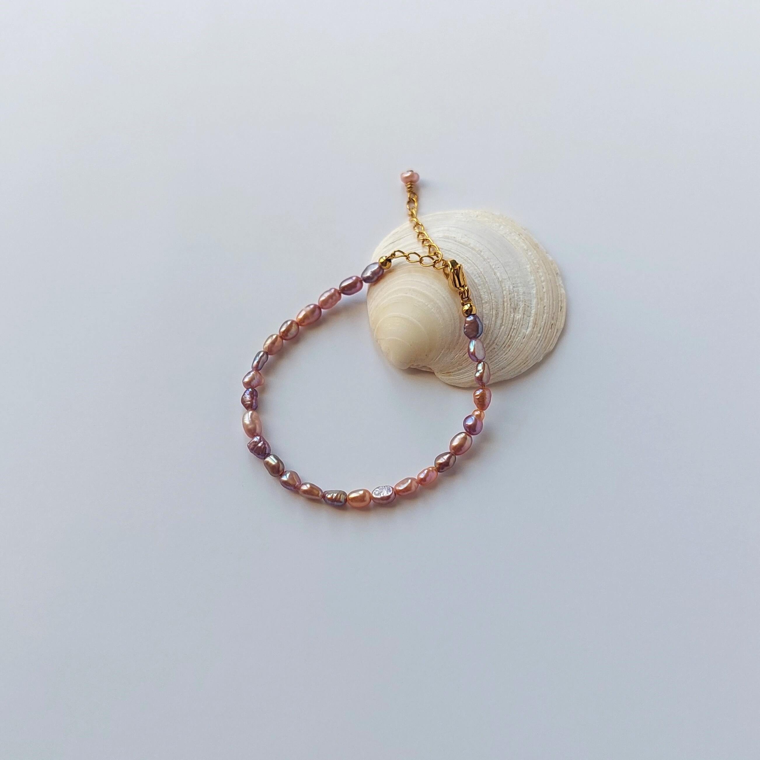Bracelet en acier inoxydable doré orné de perles d’eau douce naturelles violettes, posé sur un coquillage, style délicat et élégant, sur fond neutre. 