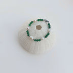 Bracelet en acier inoxydable argenté, perles d'eau douce et pierres d'aventurine verte foncée  posé sur un coquillage sur fond blanc