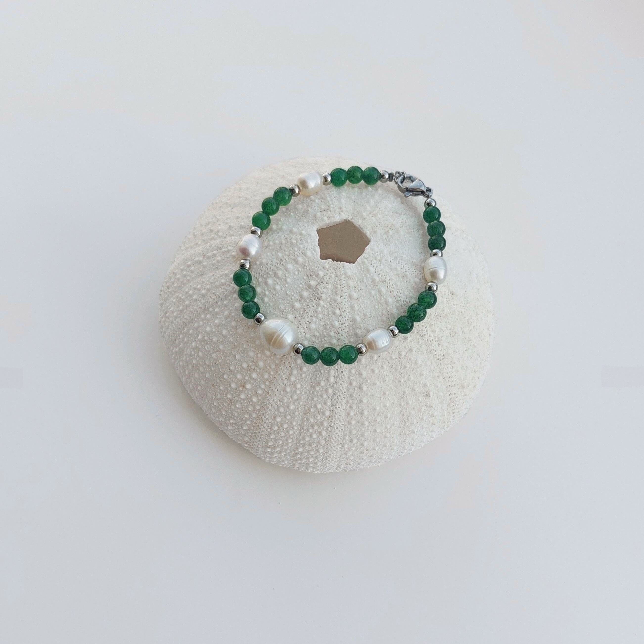 Bracelet en acier inoxydable argenté, perles d'eau douce et pierres d'aventurine verte foncée  posé sur un coquillage sur fond blanc
