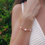 Femme portant le bracelet Galaxea composé de pierres de quartz fraise roses et de perles d'eau douce baroques entrecoupées de perles dorées en acier inoxydable, photographiée en extérieur avec un haut en crochet blanc et un fond de végétation verte.