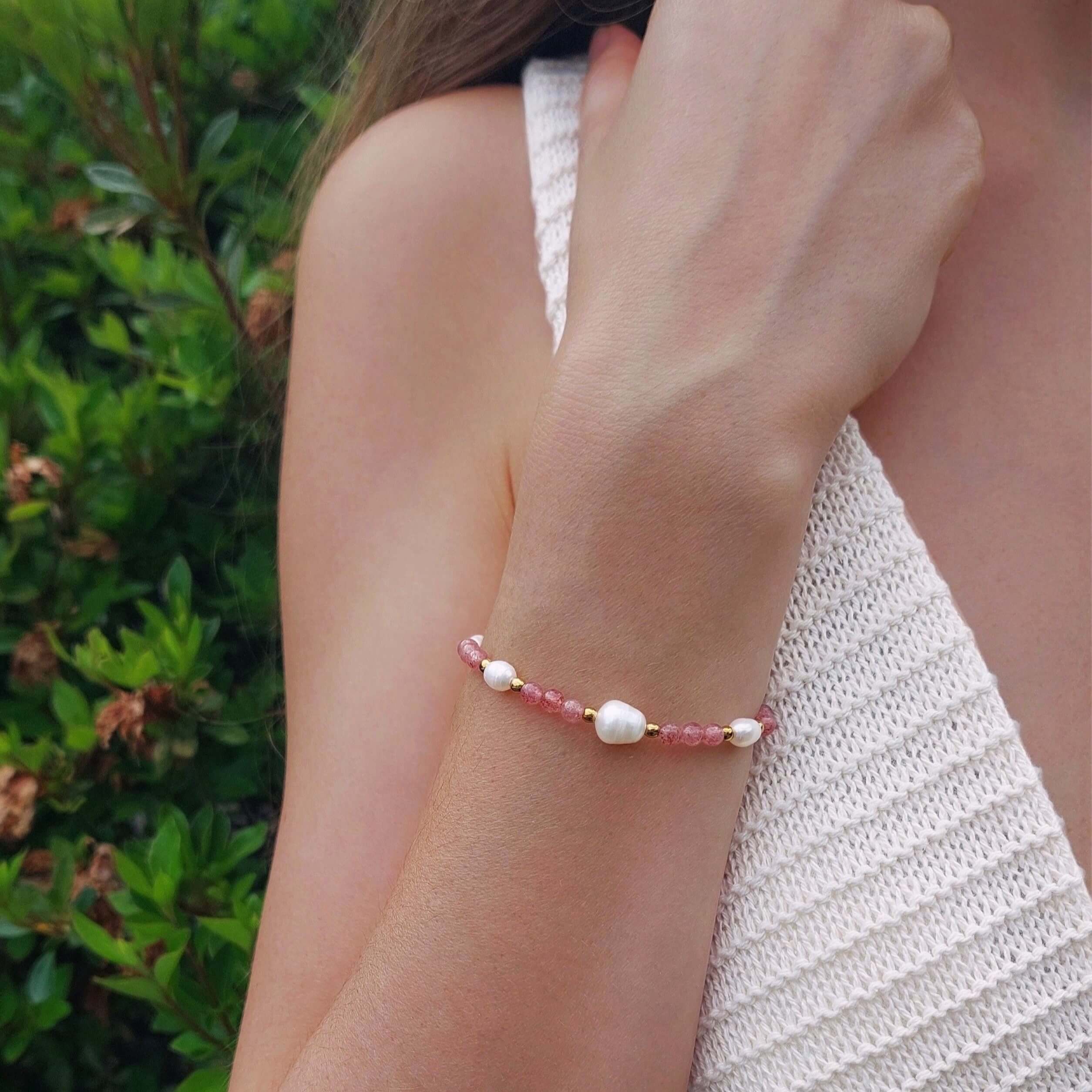 Femme portant le bracelet Galaxea composé de pierres de quartz fraise roses et de perles d'eau douce baroques entrecoupées de perles dorées en acier inoxydable, photographiée en extérieur avec un haut en crochet blanc et un fond de végétation verte.