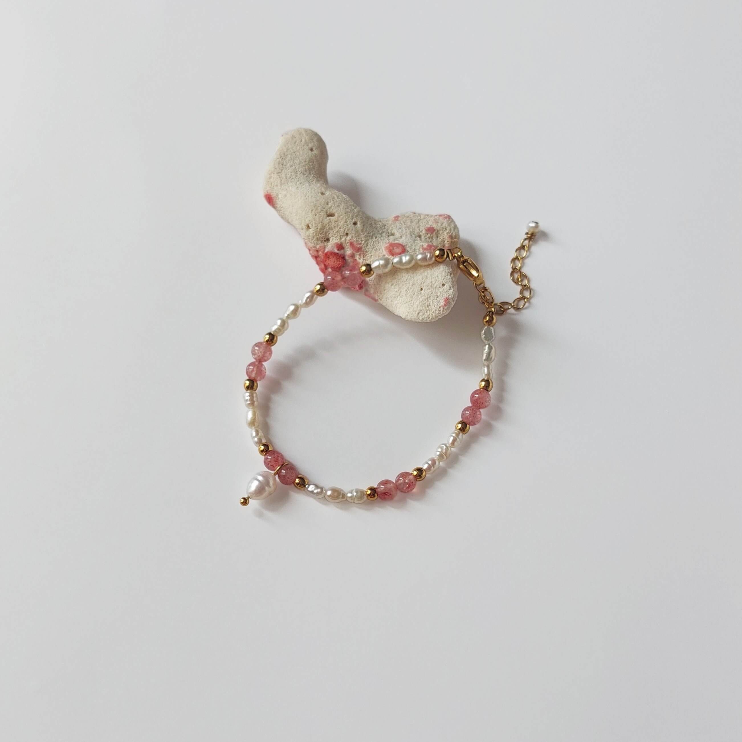 Bracelet en quartz fraise et petites perles d'eau douce baroques entrecoupées de perles dorées en acier inoxydable, posé sur un morceau de corail blanc sur fond gris clair.