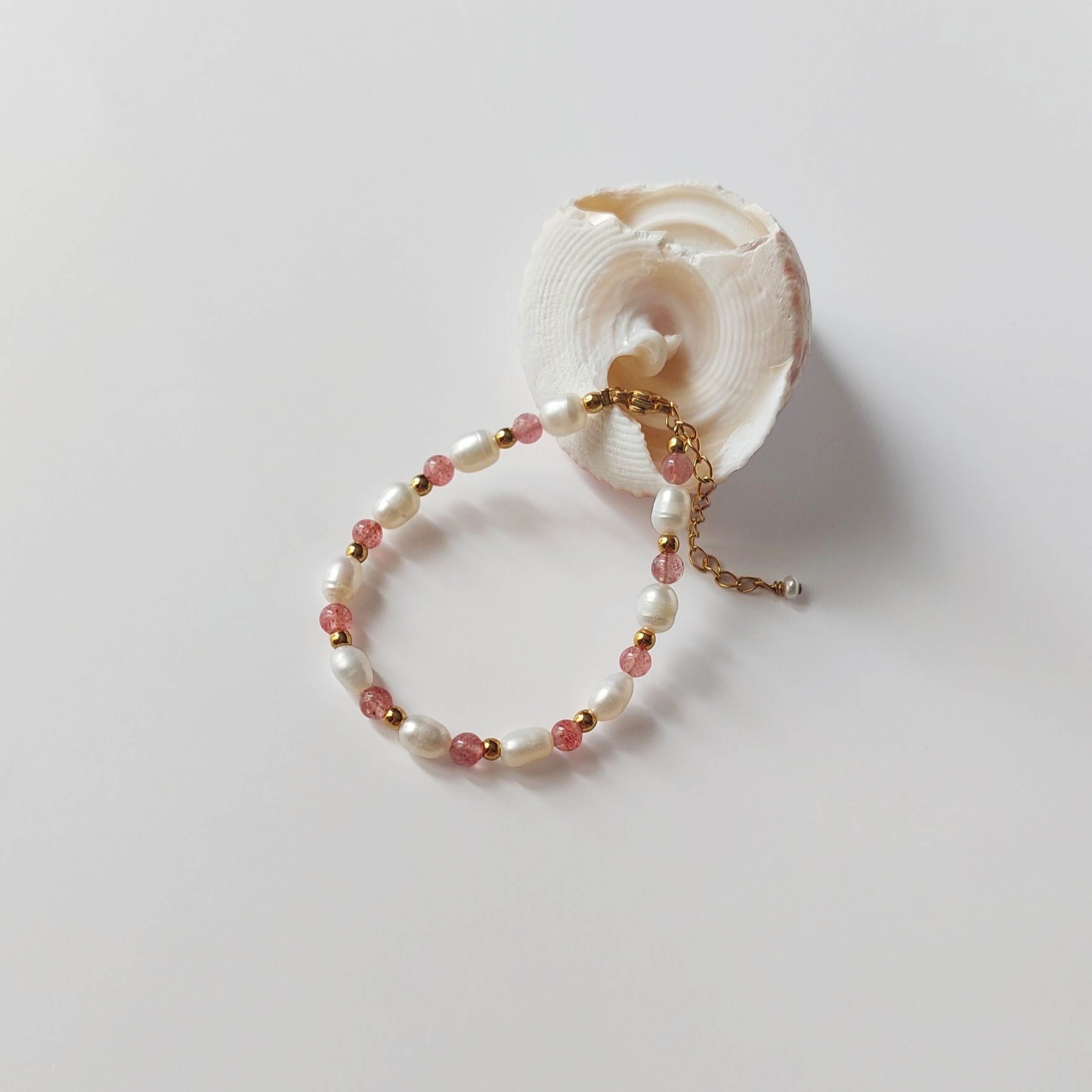 Bracelet Palythoa en perles d'eau douce baroques et pierres de quartz fraise alternées, entrecoupées de perles dorées en acier inoxydable, posé contre un coquillage blanc en spirale sur fond gris clair.