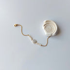 Bracelet Solaris en acier inoxydable doré avec perles d’eau douce naturelles blanches et perle centrale plate, bijou élégant et minimaliste. Posé à côté d'un coquillage et sur fond neutre.