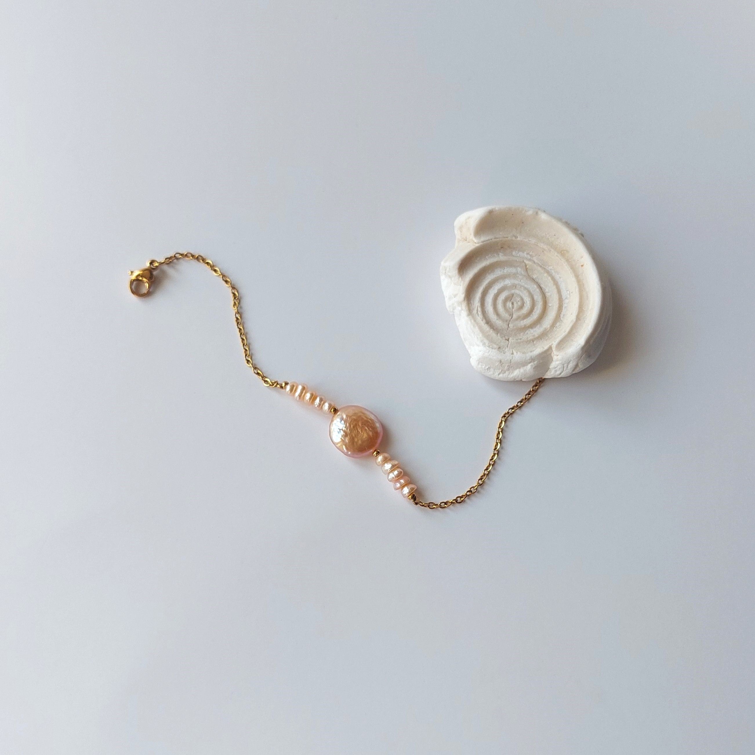 Bracelet Solaris en acier inoxydable doré avec perles d’eau douce naturelles oranges et perle centrale plate, bijou élégant et minimaliste. Posé à côté d'un coquillage et sur fond neutre.