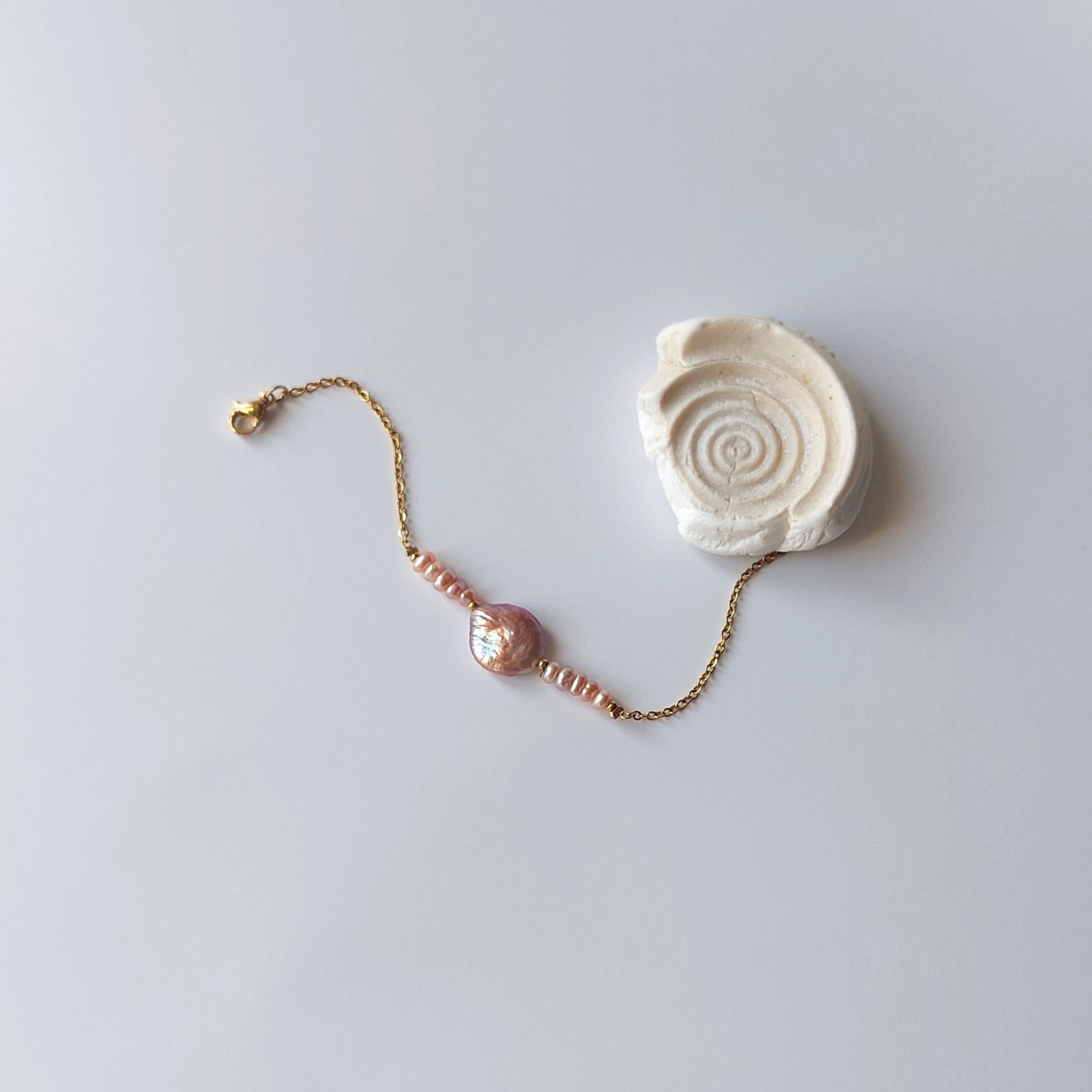 Bracelet Solaris en acier inoxydable doré avec perles d’eau douce naturelles violettes et perle centrale plate, bijou élégant et minimaliste. Posé à côté d'un coquillage et sur fond neutre.